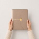 agenda de carpeta Planifesting 2027 beige croco sostenida entre las manos, broche dorado Planifesting