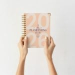 Agenda Planifesting 2026 rosa con morado sostenida con ambas manos fondo blanco