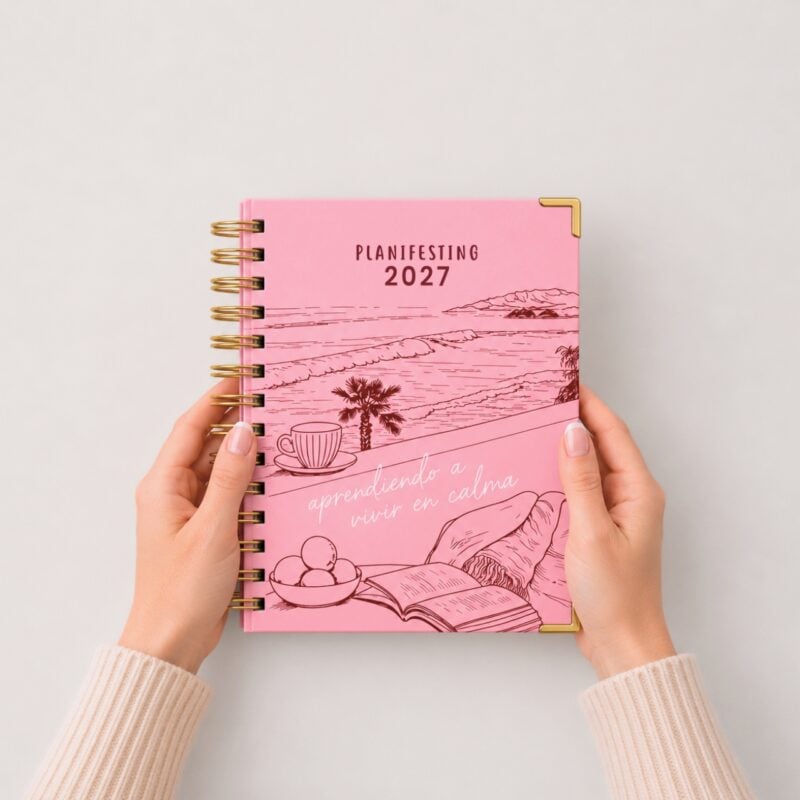 manos sosteniendo agenda de pasta dura Planifesting 2027 rosa "aprendiendo a vivir en calma" sobre fondo claro