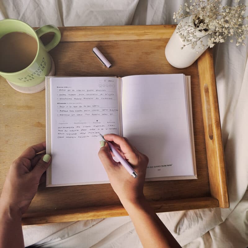 Mujer escribiendo en el Diario de Manifestación Tiempo Conmigo sobre una mesa de madera con café y flores. Diario abierto con páginas para gratitud y manifestación.