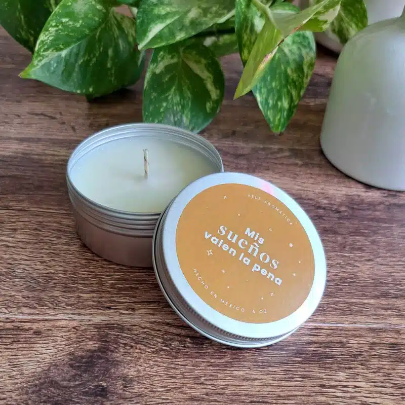 Vela aromática en lata plateada con tapa dorada que dice “Mis sueños valen la pena”, sobre mesa de madera con plantas verdes decorativas al fondo.