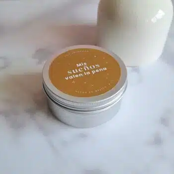 Vela aromática en lata plateada con tapa dorada que dice “Mis sueños valen la pena”, colocada sobre fondo blanco de mármol junto a un jarrón decorativo.