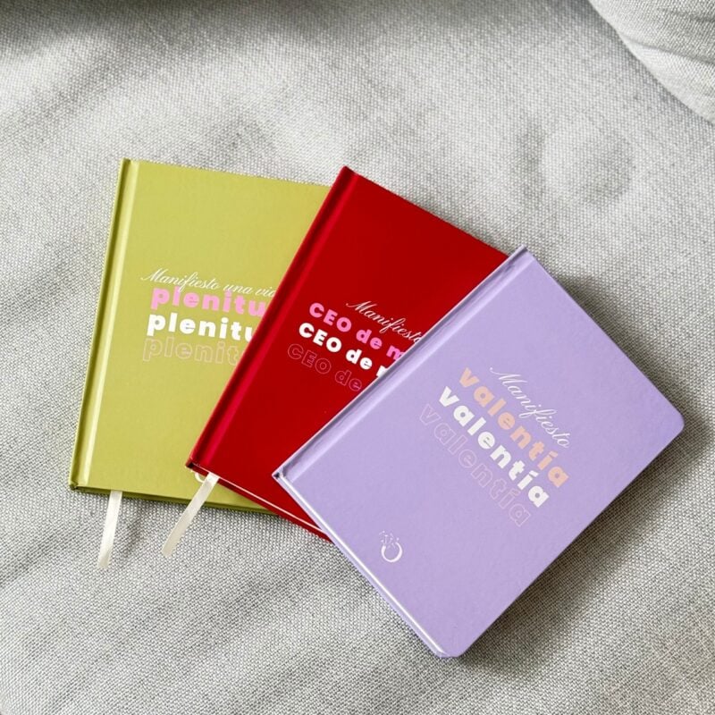 Libretas de colores morado, verde y rojo con frases inspiracionales en portada, colocadas sobre tela gris.