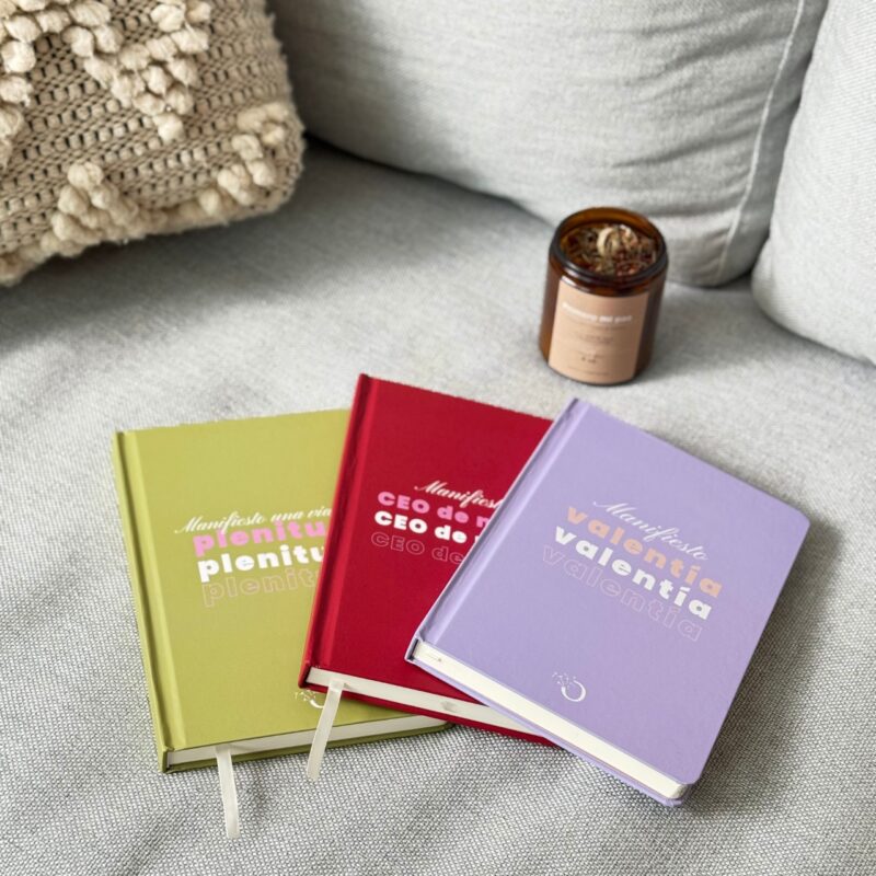 Libretas de colores morado, verde y rojo con frases inspiracionales en portada, colocadas sobre tela gris y vela