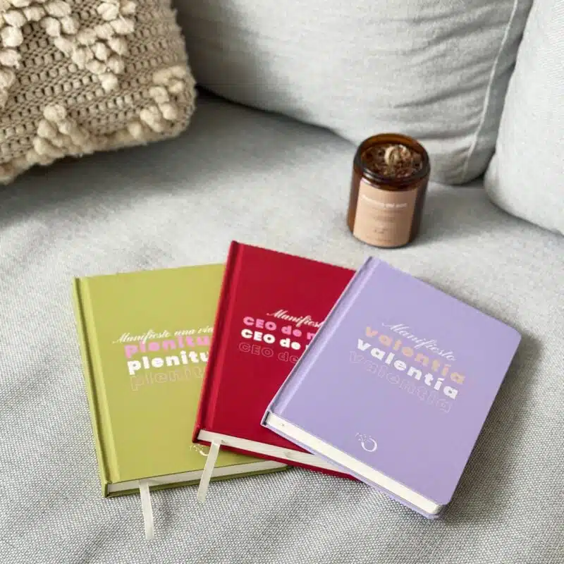 Libretas de colores morado, verde y rojo con frases inspiracionales en portada, colocadas sobre tela gris y vela