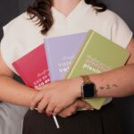 Mujer sosteniendo tres libretas 2026 de colección: libreta CEO roja, libreta Valentía morado y libreta Plenitud verde.