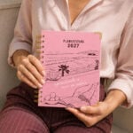 mujer sosteniendo agenda de pasta dura Planifesting 2027 rosa "aprendiendo a vivir en calma", blusa satinada rosa
